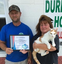Two animal shelter staff members stand outside the building, one holding an framed award and the other holding a cat.